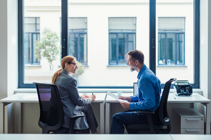 Two colleagues discuss documents at a bright windowed desk in a modern office setting.