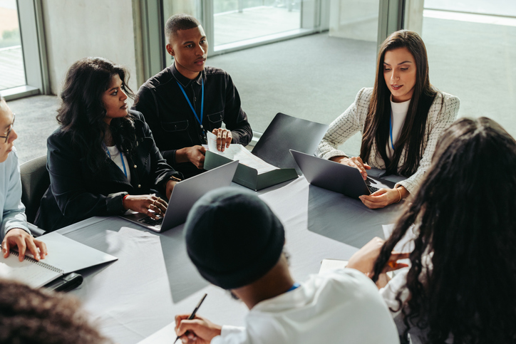 Diverse group of business people collaborating in an office setting. They engage in teamwork and discussion, working together on laptops.