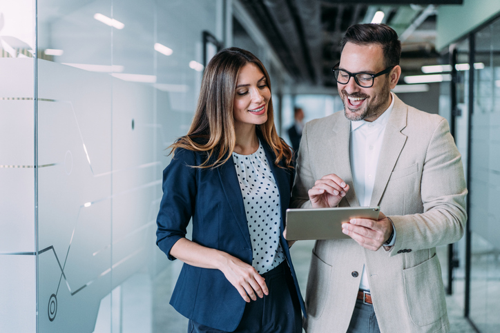 Shot of a two confident business persons talking in the work place. Two colleagues using a digital tablet while walking in a modern office. Businessman and businesswoman in meeting discussing business strategy. Business coworkers working together in the office. | How Life Sciences Marketing Operations Can Master Speed and Compliance
